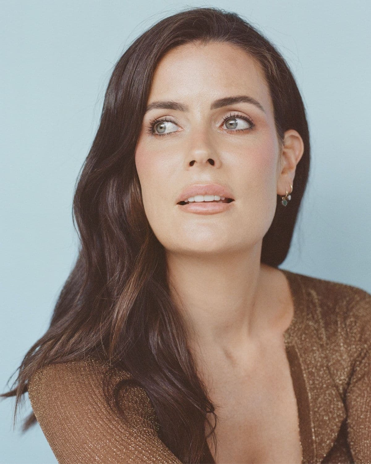 Portrait of a woman with long dark hair (likely founder Emma Lewisham) looking upward and to the side, wearing a bronze metallic top, against a pale blue backdrop