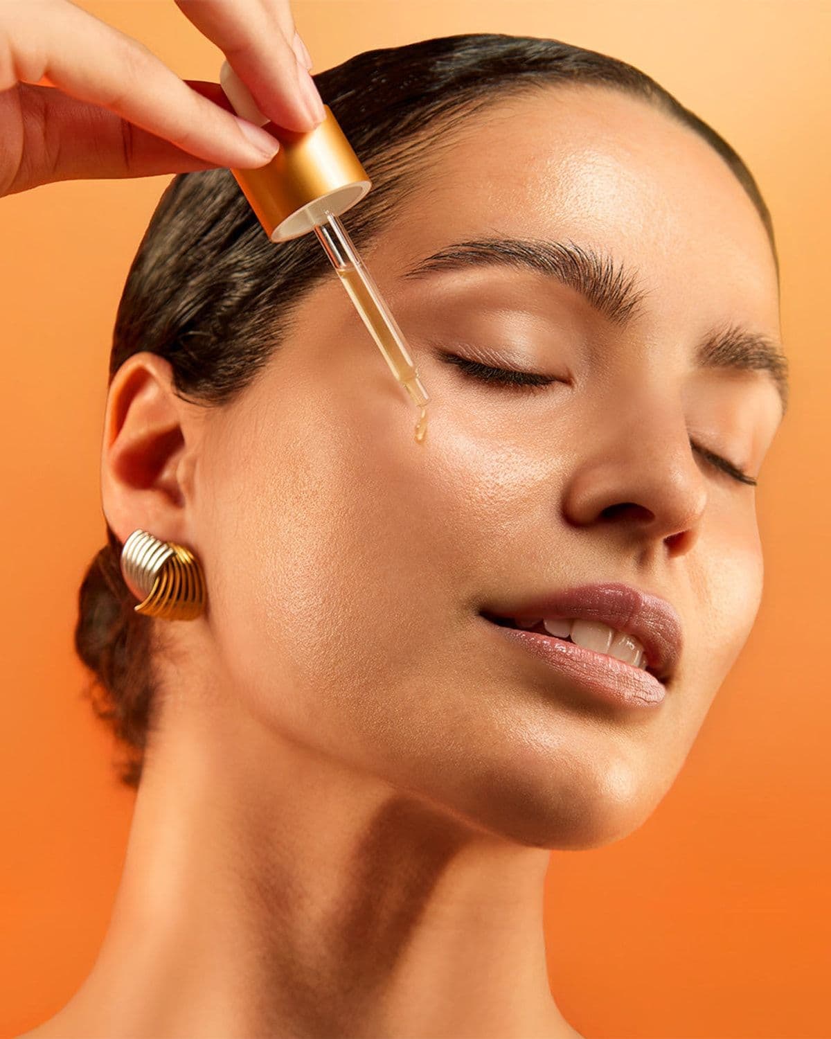 Model with wet slicked-back hair and closed eyes, applying esmi Brightening 15% Vitamin C Serum to her cheek with the dropper, warm orange campaign backdrop