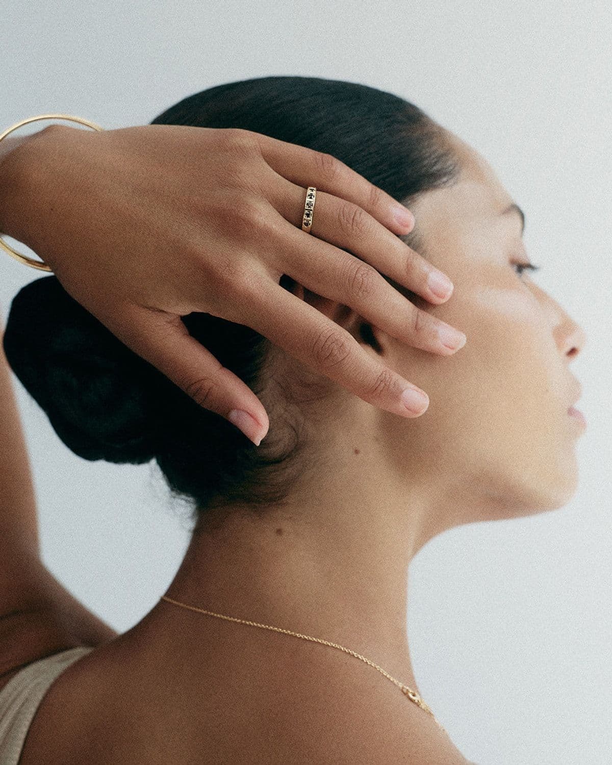 Model in profile with hand touching hair, wearing a gemstone ring and a gold bangle, soft grey backdrop