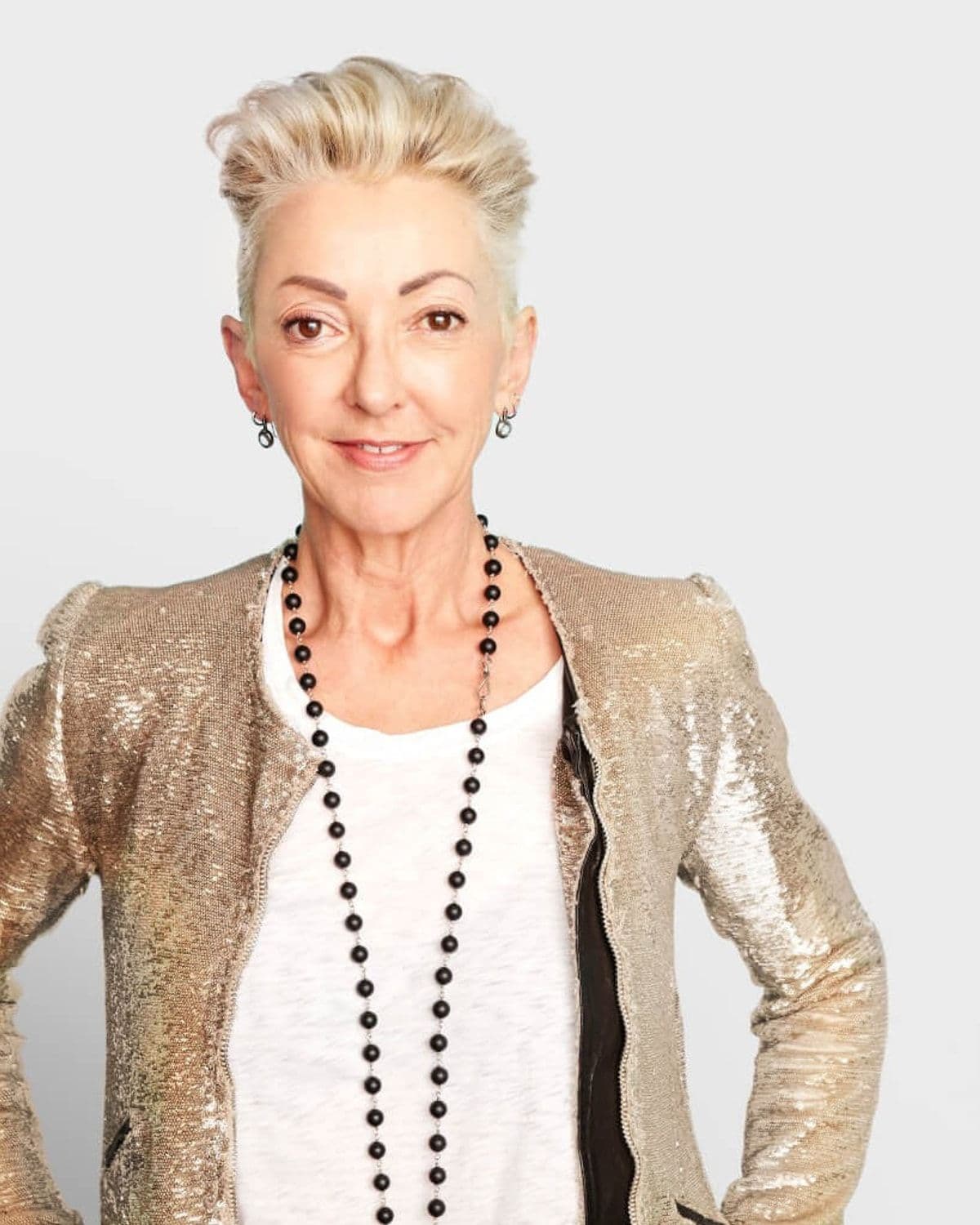 Portrait of Dermalogica founder Jane Wurwand - short blonde hair, gold sequin jacket, layered black beaded necklace - standing hands-on-hips against a plain grey background