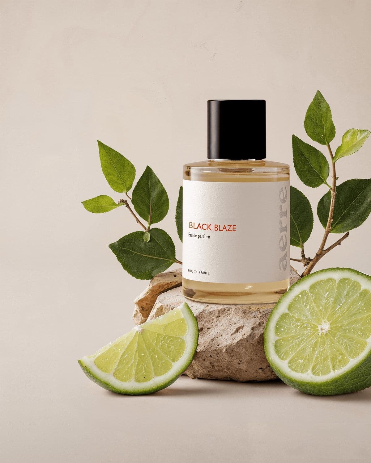 Àerre 'Black Blaze' perfume bottle staged on a stone plinth flanked by fresh green leaves and two halved limes/bergamots, cream backdrop