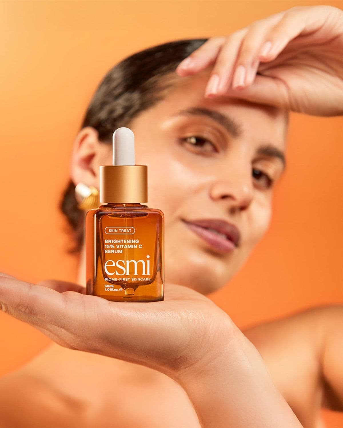 Model with wet slicked-back hair cradling an esmi Vitamin C Serum bottle in her open palm, other hand raised near her forehead, orange campaign backdrop