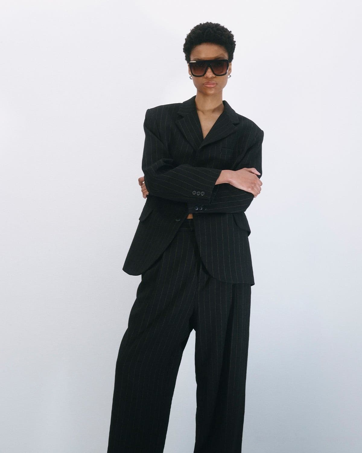 Model in an oversized black pinstripe blazer and wide-leg trousers, arms crossed, oversized sunglasses, confident editorial stance