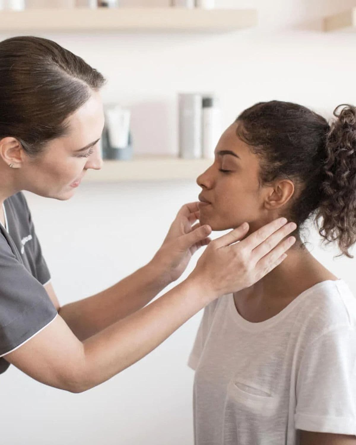 A skin therapist in a grey uniform gently examines the jawline and neck of a seated client in a bright, tiled treatment room, illustrating Dermalogica's professional skin-therapist training