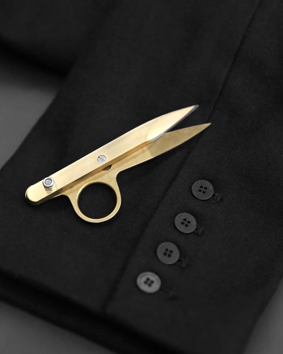 Macro editorial still-life of gold tailor's shears resting on a black wool blazer sleeve with tonal horn buttons.