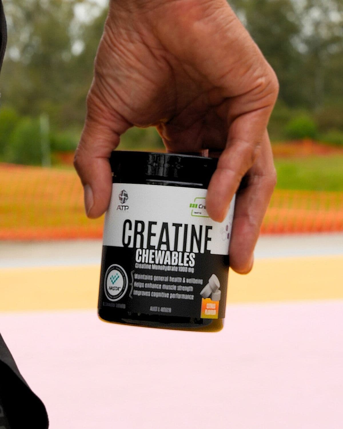 Hand holding a NOWAY! Creatine Chewables tub outdoors on a running track