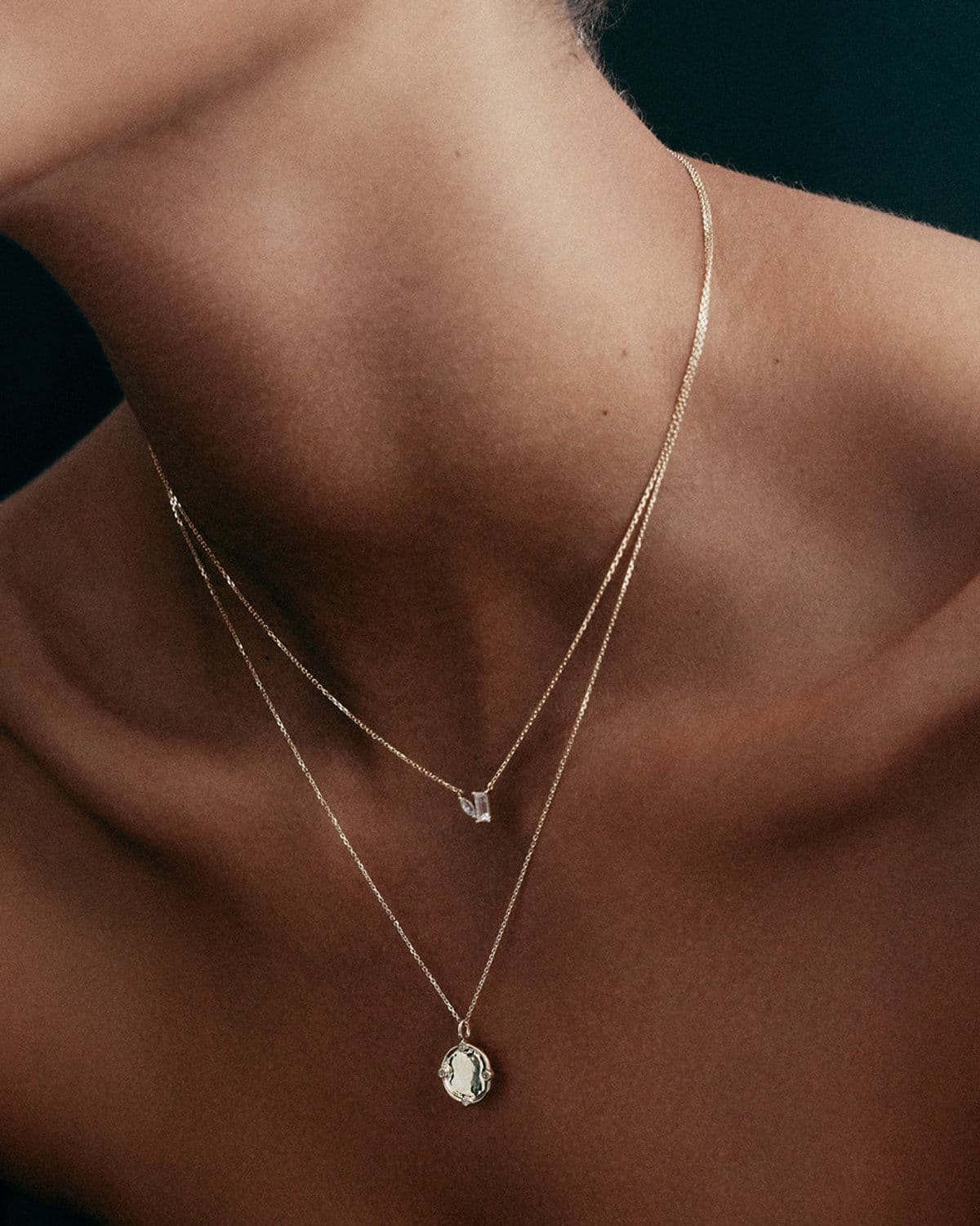 Model's bare back and shoulder in dramatic dark lighting, wearing two layered gold necklaces with pendant charms