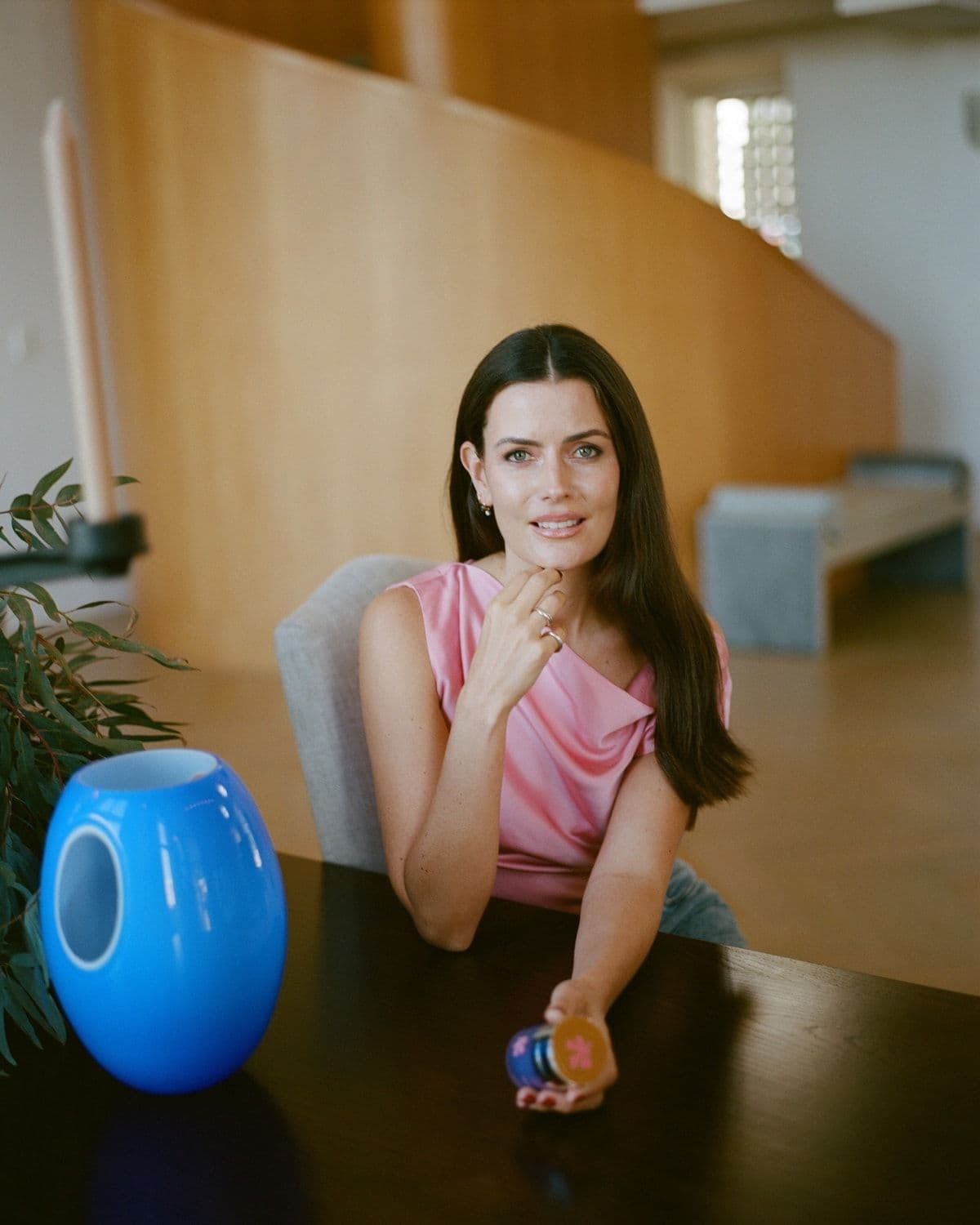Portrait of a woman with dark hair (likely founder Emma Lewisham) in a pink satin top, seated at a wooden table with a blue glass vase and eucalyptus leaves in the foreground, in a sunlit interior