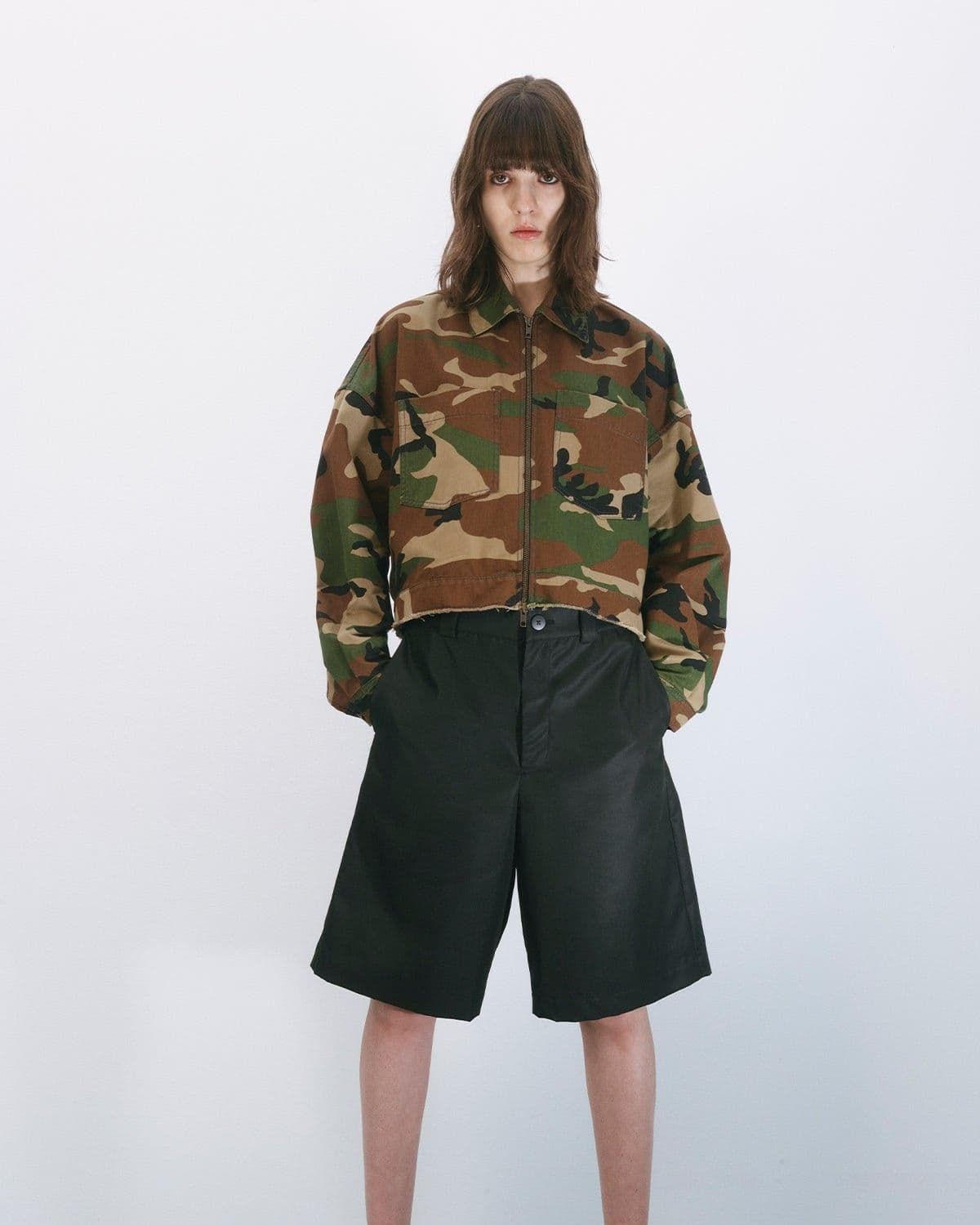Model in a cropped camo jacket and black tailored shorts, direct-to-camera gaze, minimal styled studio shot