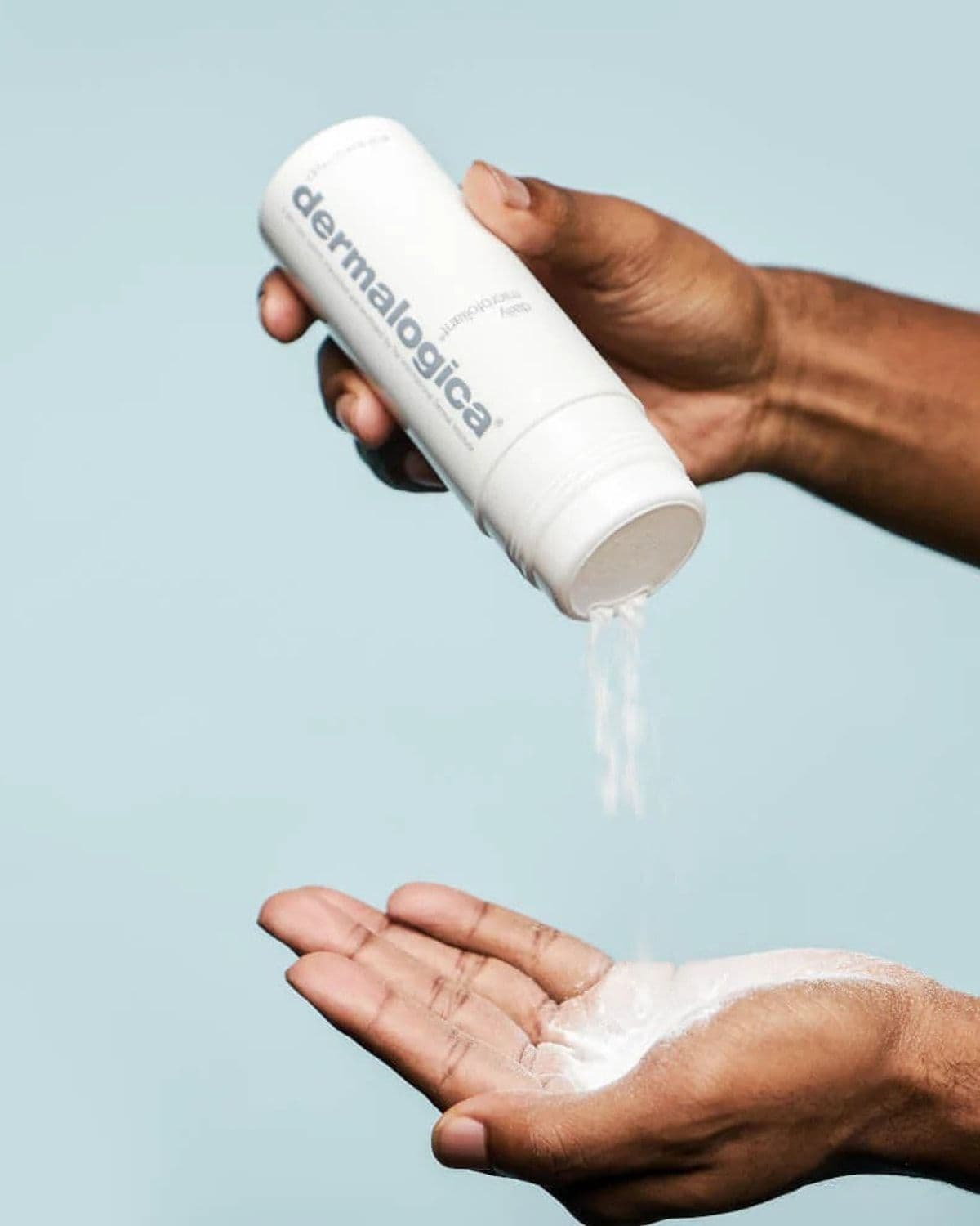 Close-up of two hands - one pouring Daily Microfoliant powder exfoliant from its bottle into the open palm of the other - against a pale teal background