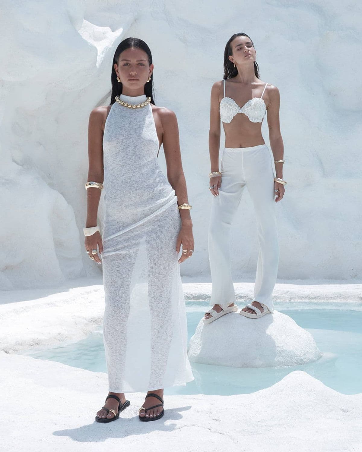 Two models in white knit and pearl-embellished outfits standing on white rock terraces beside a turquoise pool, one wearing black thong sandals