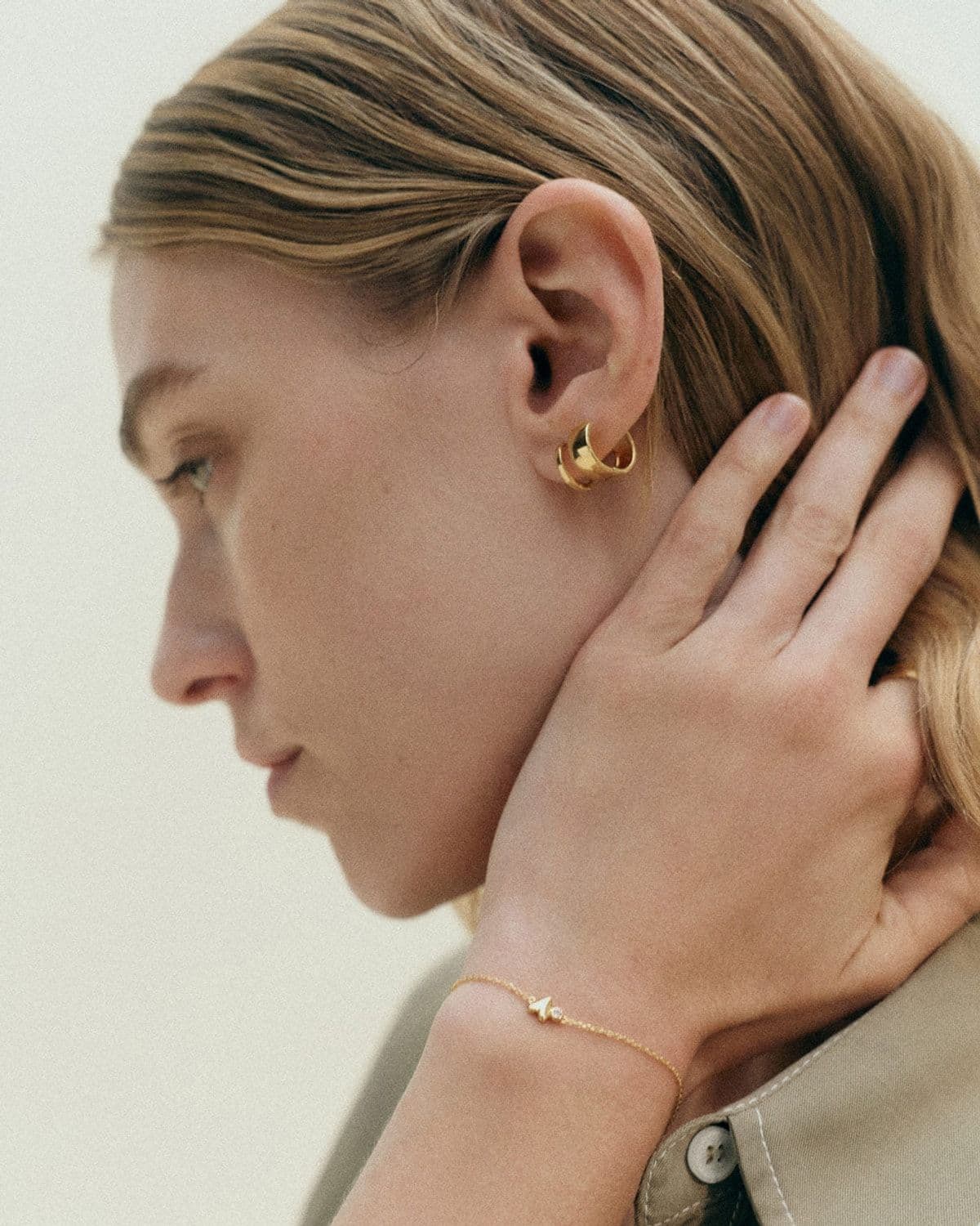 Model in profile with hand raised to hair, wearing gold hoop earrings and a delicate diamond bracelet, warm studio lighting