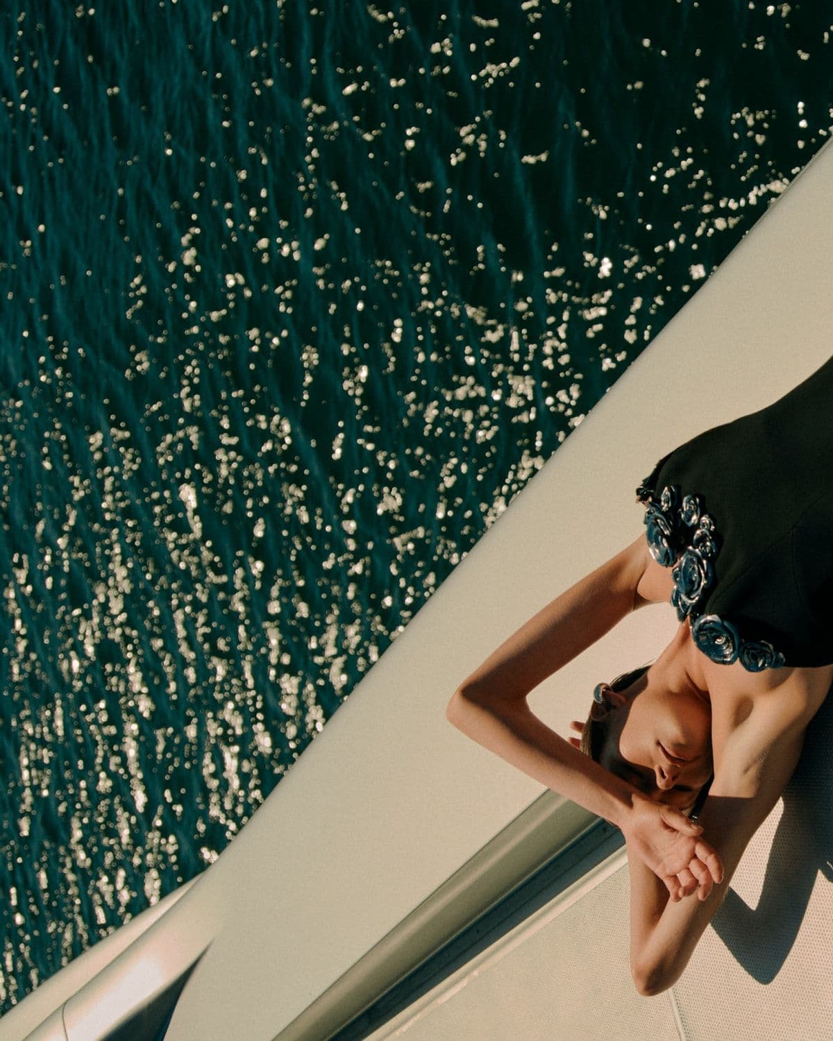 Model in black floral-appliqué gown reclining on a boat above glittering water, Pre-Fall '26 campaign