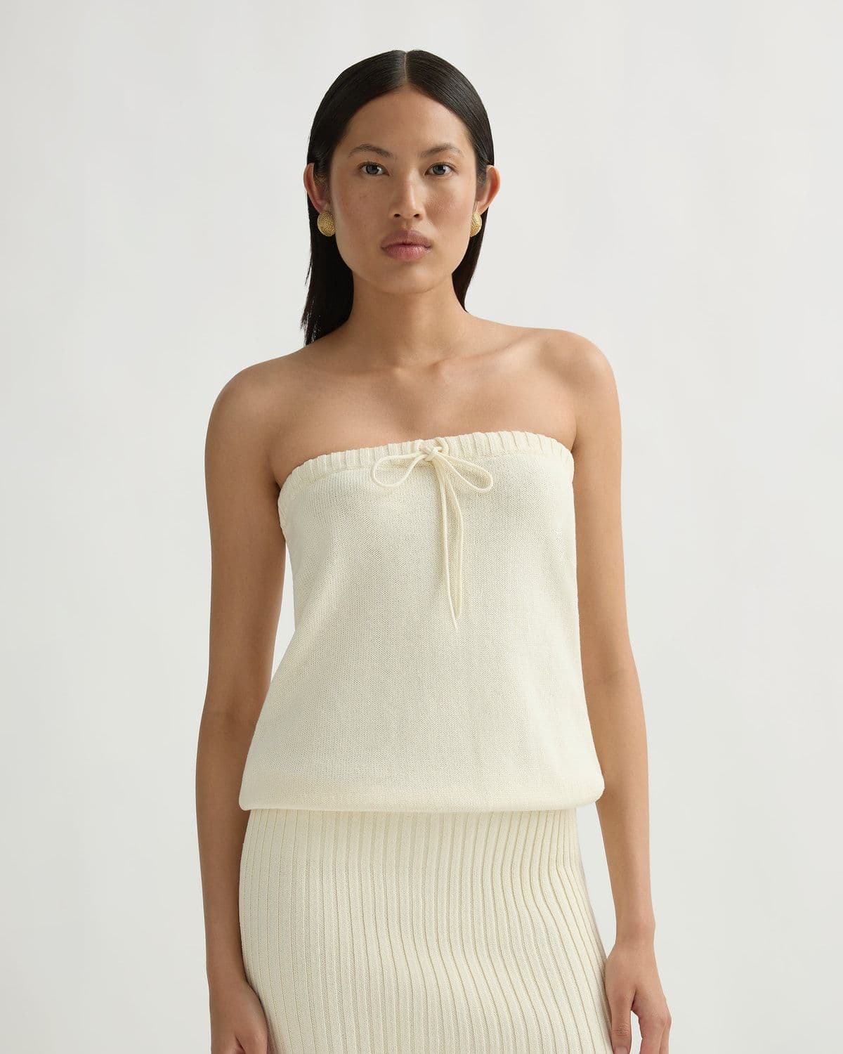 Model in an ivory open-weave crochet mini dress bending to arrange a woven basket beside a potted olive tree in a boutique interior