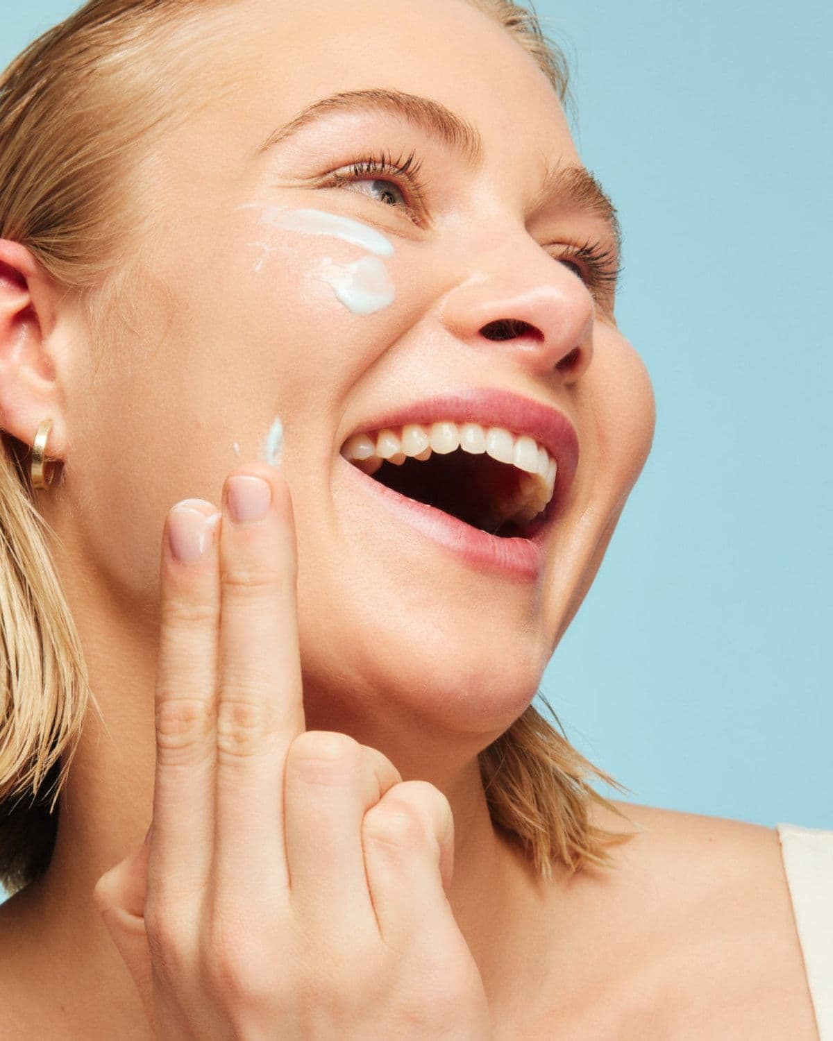 Laughing model applying esmi moisturiser to her cheek with her fingers, soft blue studio backdrop, gold hoop earrings