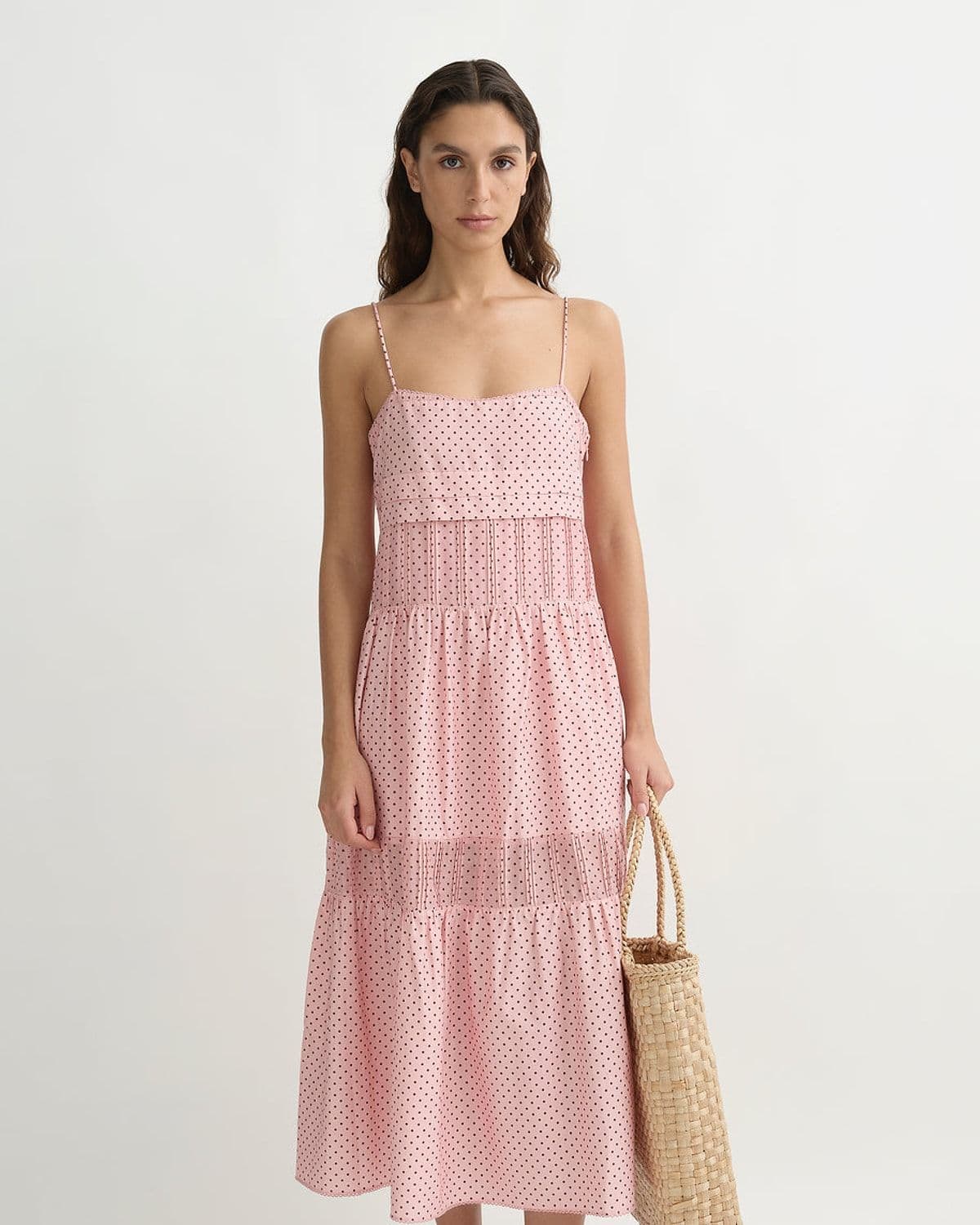 Model wearing a pink polka-dot tiered cotton-silk midi dress with spaghetti straps, carrying a woven straw tote bag, studio shot on white background