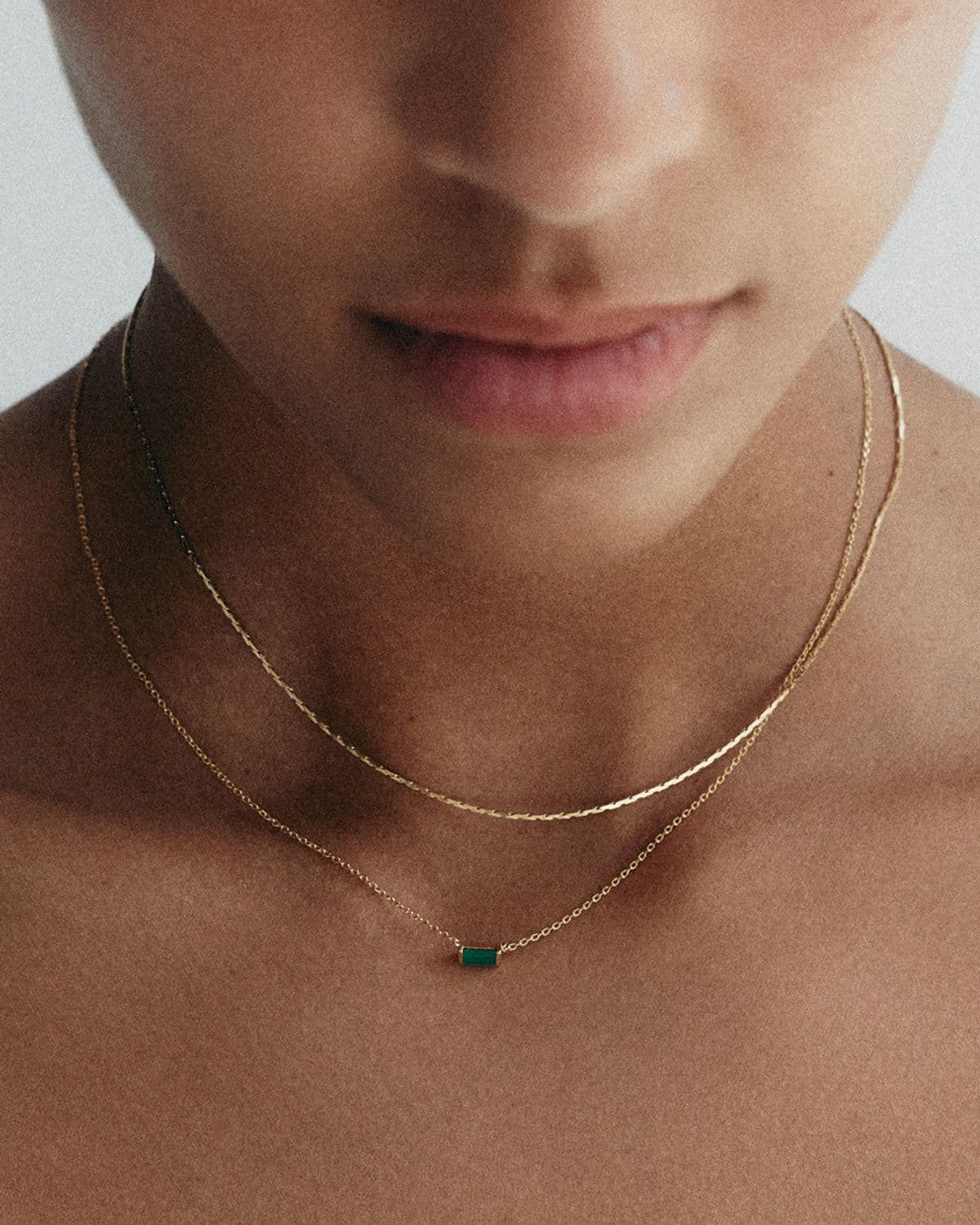 Close-up of model's collarbone and neck wearing two layered gold chain necklaces with a small emerald baguette pendant