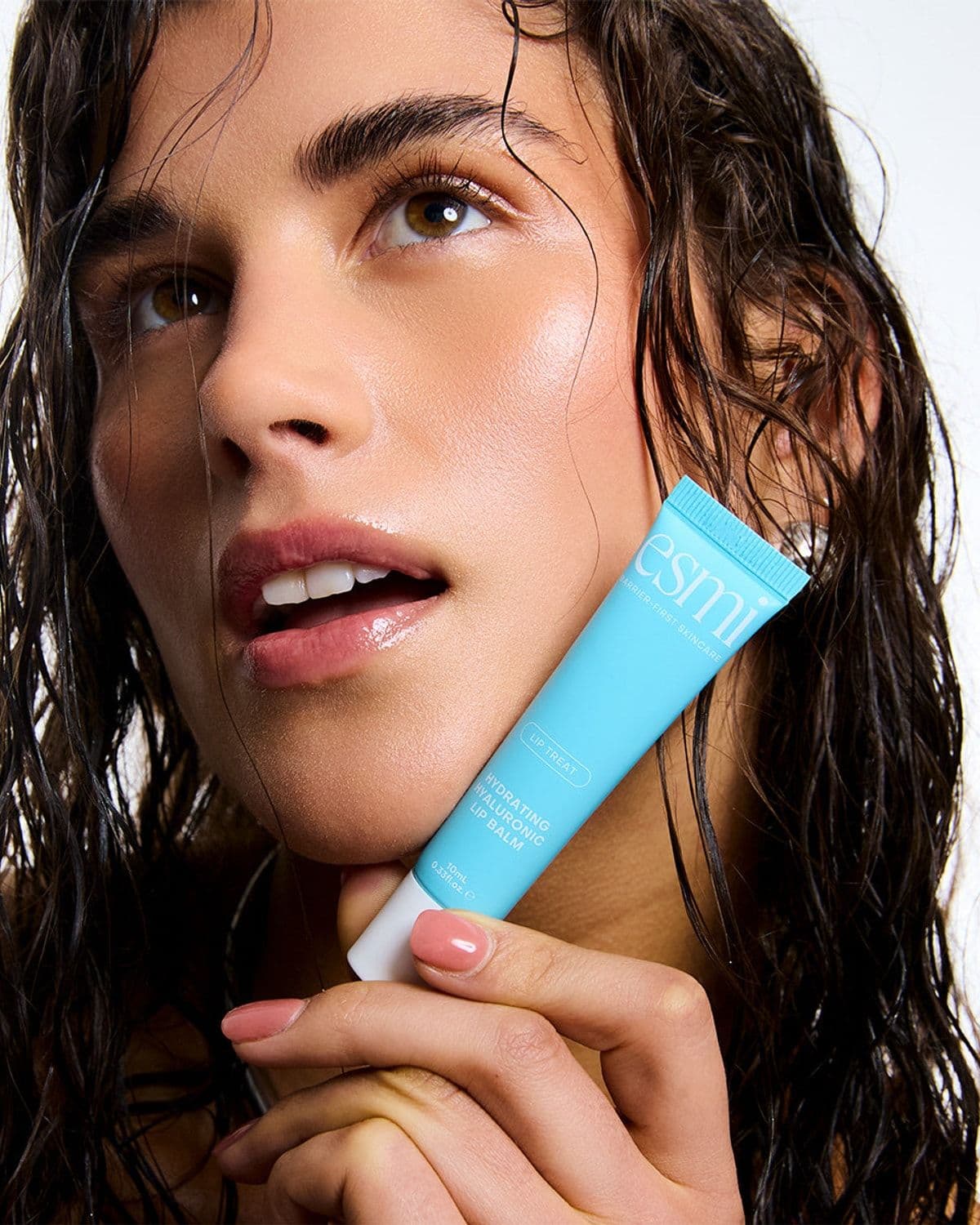 Model with wet hair looking upward, holding an esmi Hydrating Hyaluronic Lip Balm tube to her chin, bright white backdrop