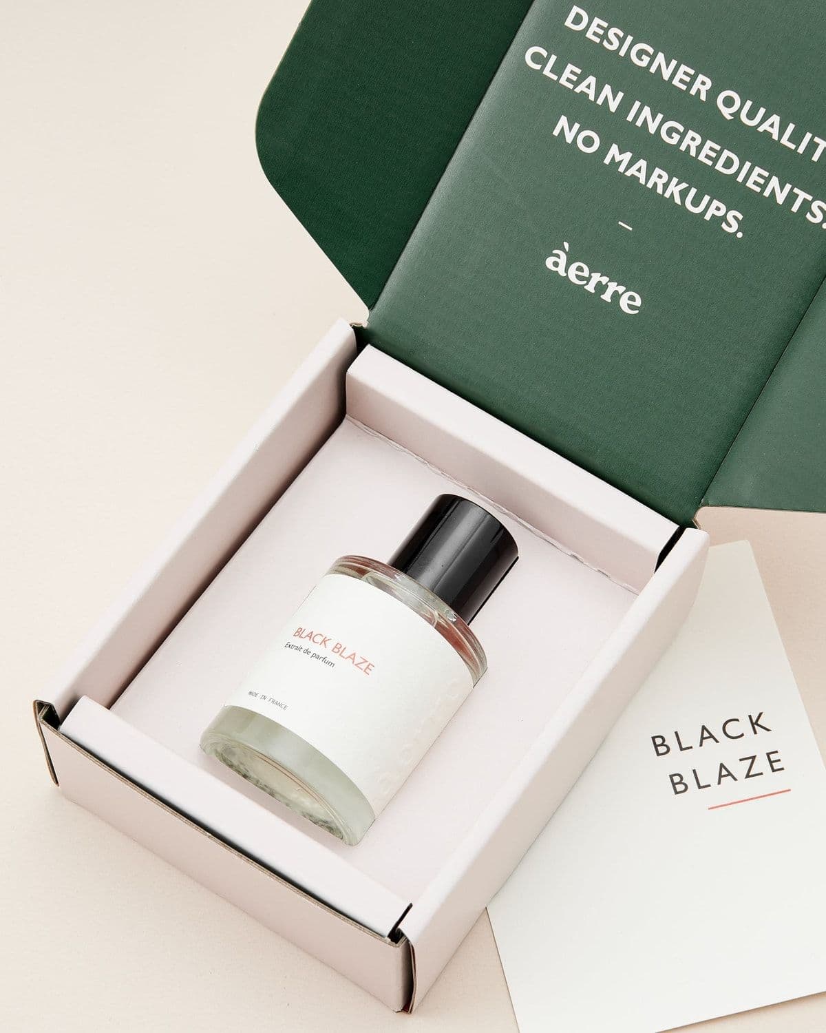 Overhead flatlay of an opened Àerre shipping box for 'Black Blaze', showing the bottle nestled in packaging next to a green insert reading 'Designer quality. Clean ingredients. No markups.' and a branded product card