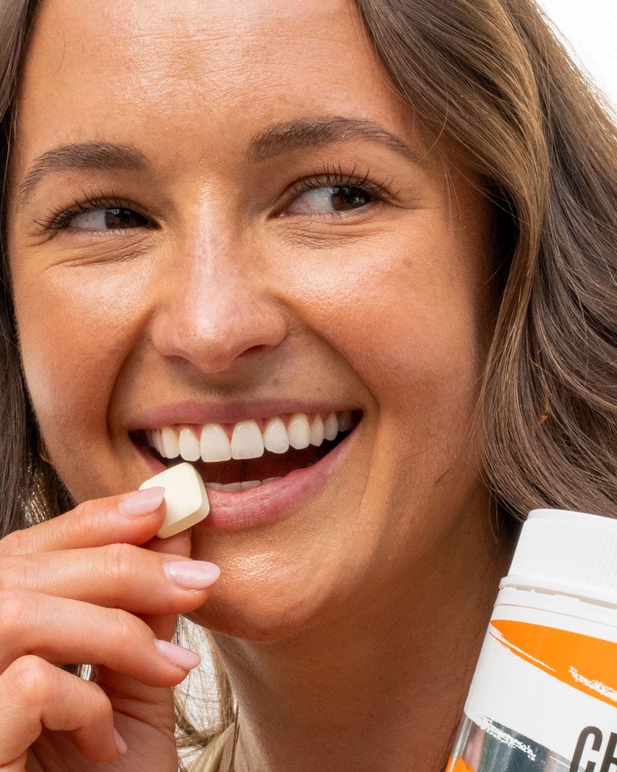 Close-up of a smiling woman biting into a NOWAY! Creatine chewable tablet, tub in foreground