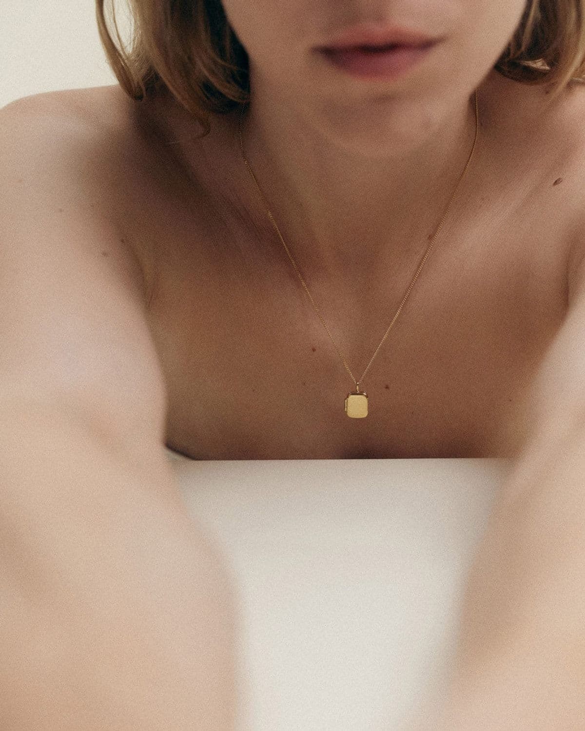 Soft-focus editorial shot of a model's bare chest and blurred face wearing a small gold locket pendant necklace, minimalist white plinth prop
