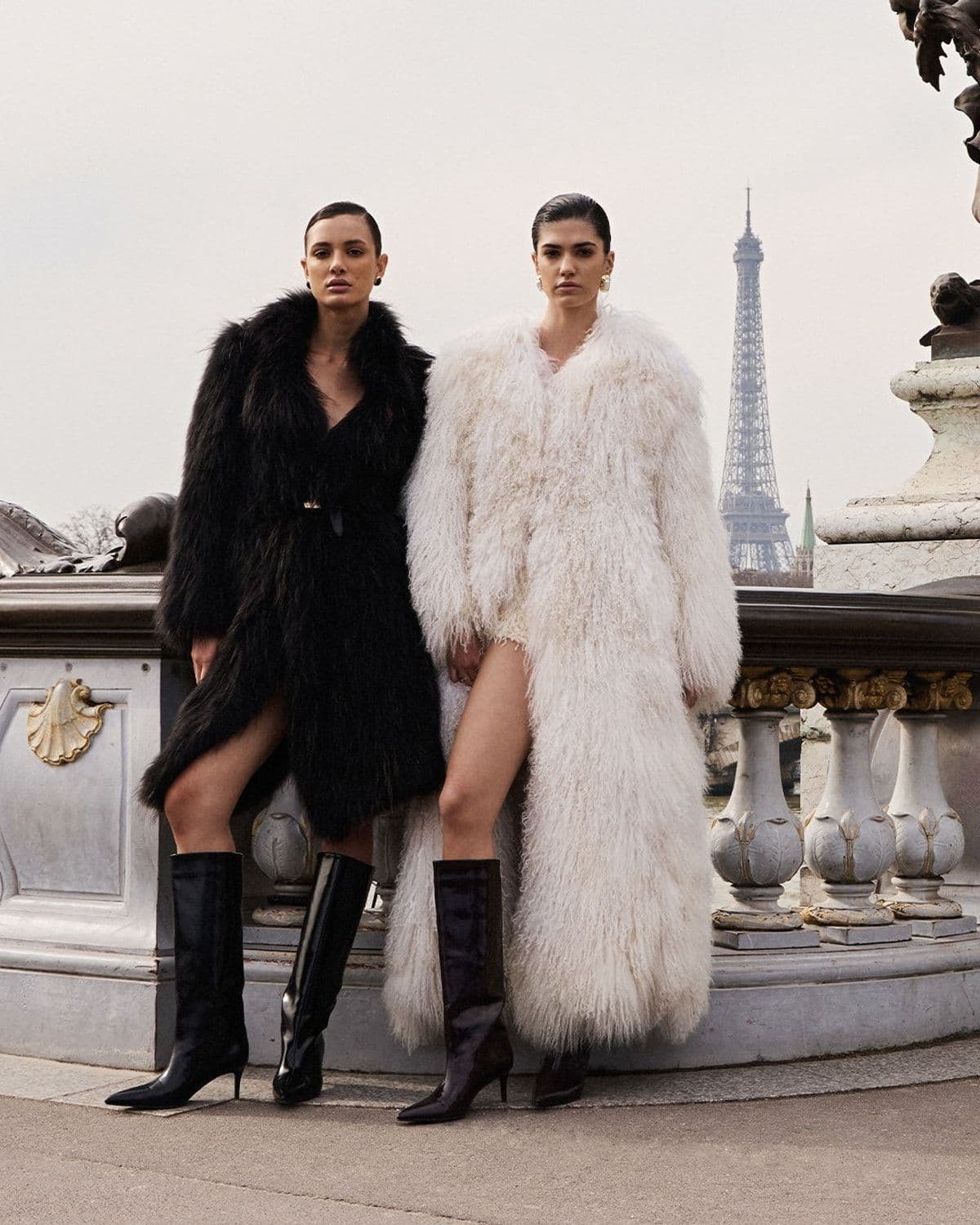 Two models in black and cream shaggy fur coats with knee-high leather boots, posed on a stone balustrade with the Eiffel Tower in the background