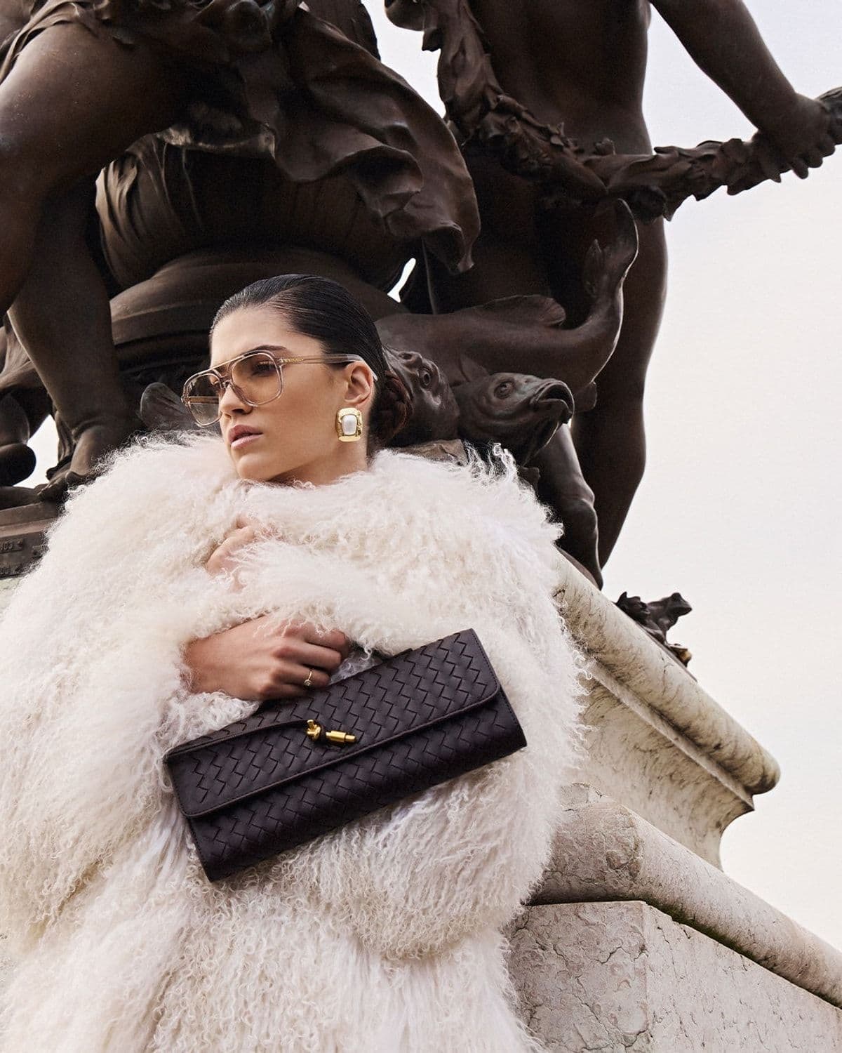 Woman in a cream shaggy fur coat and aviator sunglasses holding a dark woven leather clutch, posed against a bronze and marble monument (Fontaines de la Concorde style statue)